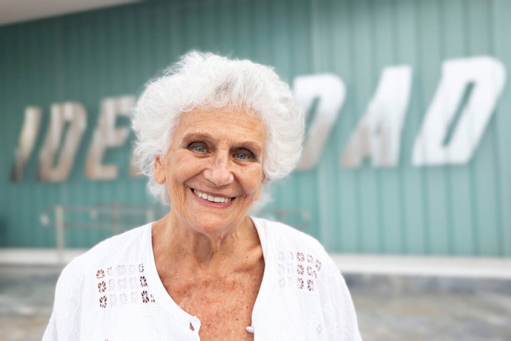 Retrato de mujer mayor sonriendo frente a fondo de pared verde con letras blancas.